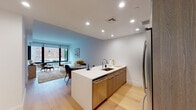 Located at 15 Bond Street in Great Neck Plaza, New York, this modern single-floor residence offers a thoughtfully designed open-concept layout with clean lines and contemporary finishes throughout. Light hardwood flooring runs seamlessly across the living areas, complementing the crisp white walls and recessed lighting overhead.

The kitchen serves as a central hub of the space, anchored by a sleek white quartz island with an undermount sink and bar seating. Warm wood cabinetry provides ample storage, while a full suite of stainless steel appliances — including a refrigerator, range, and built-in microwave — delivers both style and function. The kitchen flows effortlessly into the dining and living areas, creating a cohesive, sociable environment.

The living room is bathed in natural light through large, floor-to-ceiling windows with roller shades, offering views of the surrounding neighborhood. The bright, airy ambiance makes the space feel open and welcoming.

The bedroom is a calm retreat, featuring a large window that invites natural light and connects to the outdoors. The bathroom is finished with a light wood vanity, white quartz countertop, a large framed mirror, and a full tub-shower combination with clean tile surroundings. An in-unit washer and dryer add everyday convenience to this well-appointed residence.