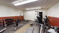 This single-floor fitness room offers a functional and well-organized workout environment suited for a range of exercise routines. The space features a drop ceiling with bright fluorescent lighting that evenly illuminates the room, creating a clean and practical atmosphere conducive to focused training.

The layout accommodates both strength training and cardio equipment. A multi-station cable machine anchors one side of the room, providing versatility for a wide variety of resistance exercises. Nearby, an adjustable weight bench and a squat/barbell rack are positioned along a wall accented with a rust-toned lower panel, offering a dedicated area for free weight workouts. Additional adjustable benches and dumbbells are distributed throughout the space. Cardio equipment, including what appears to be an elliptical trainer, is positioned toward the front.

The flooring combines a standard concrete-style surface with a central mat area, offering cushioning for floor-based exercises. The walls are finished in neutral white, with a standard door providing access. The overall ambiance is utilitarian and straightforward, prioritizing equipment accessibility and ease of movement.