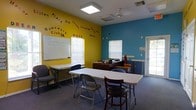 This cheerful, single-floor community room is designed to foster learning, connection, and encouragement. The space immediately draws attention with its striking two-tone color scheme — bold yellow and vivid teal walls — adorned with uplifting words and phrases such as "Hope," "Believe," "Encourage," and "Joy," as well as signage identifying it as a Homework Club and a welcoming place for gathering. A large whiteboard mounted on the yellow wall serves as a central educational tool, ideal for instruction or group activities. The room is furnished with long folding tables and a mix of chairs, offering a flexible layout that can accommodate group study sessions, tutoring, or community meetings. Natural light filters in through multiple windows fitted with blinds, complementing the overhead flush-mount and ceiling fan lighting fixtures. A workstation or desk area is visible toward the back of the room, providing a dedicated space for instructors or organizers. The carpeted floor adds warmth to the space, while a glass-panel door offers easy access. The overall ambiance is energetic yet welcoming, clearly purpose-built to motivate and support the community members it serves.