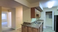 This single-story space centers around a practical and well-appointed kitchen that serves as a focal point of the interior. The kitchen features warm-toned wood cabinetry, both upper and lower, providing ample storage throughout. The countertops display a dark, stone-style laminate surface that complements the cabinetry's natural hue. A double stainless steel sink is integrated into the counter, accompanied by a standard faucet fixture, adding a clean, utilitarian element to the workspace.

The kitchen layout incorporates a peninsula-style counter configuration, creating a natural division between the cooking area and the adjoining spaces. Upper cabinetry is mounted beneath a soffit, maximizing vertical storage while maintaining a structured, finished appearance overhead. A textured ceiling treatment is visible throughout, consistent with the overall construction style.

The flooring transitions between tile and a contrasting material in the adjacent area, suggesting distinct functional zones within the open layout. Neutral light green walls provide a subdued backdrop throughout. The space opens into a connected room visible through a wide doorway, reinforcing the open and accessible flow of the single-floor layout. The overall ambiance is practical and move-in ready.