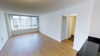 This single-floor unit features an open and airy living area finished with light-toned laminate hardwood flooring that extends throughout the main space. The room is illuminated by a large window dressed with vertical blinds, allowing natural light to fill the interior while offering views of the surrounding area. White walls and white baseboards contribute to a clean, minimalist aesthetic throughout. A textured ceiling adds a subtle architectural detail overhead. A baseboard heater runs beneath the window, providing a practical heating solution. Toward the rear of the living area, an open doorway leads to additional rooms, revealing a hallway with what appears to be a closet fitted with a hanging rod — ideal for storage. The transition between spaces is defined by clean white door frames and trim, maintaining a cohesive look. The overall layout is straightforward and functional, presenting a well-lit, move-in-ready space with a neutral palette that easily accommodates a variety of furnishing styles.