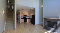 This two-story space is defined by its dramatic ceiling height, which creates an airy, open atmosphere throughout the main living area. Light hardwood flooring extends across the main level, lending warmth and continuity to the space.

The kitchen is accessible through a wide architectural opening and is equipped with black appliances, including a refrigerator, electric range, and built-in microwave. Light-toned cabinetry lines the walls, complemented by dark countertops that provide a clean contrast. Track lighting mounted to the ceiling ensures the kitchen is well-illuminated. A hallway visible beyond the kitchen suggests additional rooms or storage further into the unit.

The living area benefits from the soaring two-story ceiling, amplifying the sense of scale. A tiled fireplace with a light wood mantel is positioned along one wall, serving as a notable architectural feature and a natural focal point for the room. The neutral, muted wall tones throughout complement the flooring and contribute to a calm, cohesive aesthetic. The overall design balances modern functionality with an open, inviting ambiance.