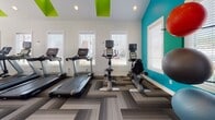 Located at 961 Marcus Dr in Newport News, Virginia, this fitness center is a thoughtfully designed single-floor facility that offers a comprehensive workout environment. The space is defined by its striking vaulted ceilings, which soar to an impressive peak and are accentuated by bold teal accent walls and vivid lime-green ceiling details, giving the gym a modern, energetic ambiance. Ceiling fans are mounted throughout to ensure comfortable airflow during workouts.

The flooring features a geometric herringbone-patterned carpet tile in neutral gray tones, adding visual interest while providing a practical surface underfoot. Full-length mirrors line one wall, enhancing the sense of openness and allowing users to monitor their form during exercise.

The cardio zone is well-appointed with multiple treadmills, stationary upright bikes, and a recumbent bike, all positioned near large windows fitted with white blinds that invite abundant natural light. The strength training area offers a diverse selection of equipment, including a multi-station cable machine, a Matrix power rack with a bench, a dedicated free weights section with a dumbbell rack, and stability and medicine balls neatly organized along the wall. Recessed lighting throughout ensures the space remains bright and well-illuminated at all times. This fitness center provides a complete and inviting environment suited for a wide range of workout routines.