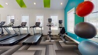 Located at 961 Marcus Dr in Newport News, Virginia, this fitness center is a thoughtfully designed single-floor facility that offers a comprehensive workout environment. The space is defined by its striking vaulted ceilings, which soar to an impressive peak and are accentuated by bold teal accent walls and vivid lime-green ceiling details, giving the gym a modern, energetic ambiance. Ceiling fans are mounted throughout to ensure comfortable airflow during workouts.

The flooring features a geometric herringbone-patterned carpet tile in neutral gray tones, adding visual interest while providing a practical surface underfoot. Full-length mirrors line one wall, enhancing the sense of openness and allowing users to monitor their form during exercise.

The cardio zone is well-appointed with multiple treadmills, stationary upright bikes, and a recumbent bike, all positioned near large windows fitted with white blinds that invite abundant natural light. The strength training area offers a diverse selection of equipment, including a multi-station cable machine, a Matrix power rack with a bench, a dedicated free weights section with a dumbbell rack, and stability and medicine balls neatly organized along the wall. Recessed lighting throughout ensures the space remains bright and well-illuminated at all times. This fitness center provides a complete and inviting environment suited for a wide range of workout routines.