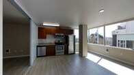 This single-floor space combines functional design with a welcoming, light-filled atmosphere. The open-plan layout seamlessly connects the kitchen and living areas, creating a cohesive and versatile living environment.

The kitchen is well-appointed with rich, warm-toned wooden cabinetry providing ample storage, complemented by granite-style countertops and a stainless steel appliance suite that includes a refrigerator, gas range, built-in microwave, and dishwasher. A dedicated sink station adds to the kitchen's practicality.

Flowing directly from the kitchen, the living area is defined by wide-plank, light-toned hardwood flooring that runs throughout the space, lending a clean and modern aesthetic. A structural column subtly divides the two zones while preserving the open feel of the floor plan.

The standout feature of the living area is the expansive floor-to-ceiling windows along the exterior wall, which flood the space with abundant natural light and frame views of the surrounding neighborhood. Recessed lighting and under-cabinet fixtures supplement the natural light, ensuring the space remains bright at all times.

The smooth white ceiling and neutral wall tones create a calm, airy backdrop that enhances the overall sense of openness throughout this thoughtfully designed space.
