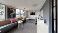 This thoughtfully designed single-floor space combines a welcoming lobby, a comfortable lounge, and a functional business center into one cohesive environment. The entry area features a combination of large-format tile and patterned carpet flooring, with linear LED lighting overhead and generous windows that frame views of the surrounding exterior. Built-in mailboxes and a front desk area anchor the reception zone, lending the space an organized and professional feel.

The lounge area exudes a relaxed yet refined atmosphere, furnished with a sleek, low-profile sofa accented by vibrant throw pillows in yellow, pink, and patterned fabrics. A pair of latticed side tables and globe pendant lights suspended from a brass-finished bar fixture add a curated, contemporary touch. Bold, colorful artwork — including a striking question mark canvas — enlivens the neutral gray walls, while large windows flood the seating area with natural light and outdoor views.

Adjacent to the lounge, the business center is defined by a whitewashed wood plank accent wall that provides a warm, textural contrast to the deep navy feature wall. A wall-mounted flat-screen TV, two computer workstations with ergonomic yellow-green task chairs, and a shared printer offer practical amenities. The overall ambiance is modern and inviting, balancing productivity with comfort throughout the space.