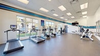 Located at 100 Reflections Circle in Casselberry, Florida, this fitness center is a single-floor facility thoughtfully divided into two dedicated exercise rooms connected by a hallway. The space is designed to support a comprehensive range of workout routines, from cardio to strength and functional training.

The primary cardio room is bright and open, lined with large windows that flood the space with natural light and offer views of the surrounding grounds. The room features a row of treadmills, elliptical trainers, and stationary bikes positioned along the windowed wall, complemented by a wall-mounted flat-screen television. A distinctive blue and gray striped accent band runs along the upper walls, giving the space a clean, energetic aesthetic. Durable rubber flooring and a drop-panel ceiling with recessed lighting complete the polished look.

The second exercise room is dedicated to strength and functional training. It is outfitted with a multi-station cable and functional trainer rig in bold orange, a power rack, weight benches, a punching bag, medicine balls, dumbbells, battle ropes, and a heavy tire for high-intensity training. Mirrored walls expand the sense of space and aid in form correction. A separate area within the room accommodates cycling bikes and open floor space for group or individual training sessions. Together, the two rooms offer a versatile, well-appointed fitness environment.
