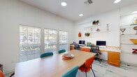 Located at 5500 Sampson St in Houston, TX, this single-floor office space is thoughtfully designed to support both collaborative work and individual productivity. The room is anchored by a large central conference table surrounded by a mix of teal and orange ergonomic chairs, making it well-suited for meetings or group work sessions. Along one wall, a built-in wooden workstation provides dedicated computer terminals with task chairs, complemented by floating display shelves adorned with decorative art glass, sculptures, and colorful accent pieces that lend the space a creative, contemporary character. A patterned carpet in light tones with teal accents adds visual interest underfoot. Multiple large windows fitted with horizontal blinds line two walls, flooding the space with natural light while offering views of the surrounding greenery. Vibrant, large-format artwork displayed on the walls further enhances the energetic ambiance. A cozy seating nook with patterned armchairs provides an informal lounge area, and a glass-paned interior door connects to an adjacent space. Recessed ceiling lighting ensures consistent illumination throughout. The overall atmosphere is bright, modern, and professionally inviting.