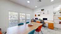 Located at 5500 Sampson St in Houston, TX, this single-floor office space is thoughtfully designed to support both collaborative work and individual productivity. The room is anchored by a large central conference table surrounded by a mix of teal and orange ergonomic chairs, making it well-suited for meetings or group work sessions. Along one wall, a built-in wooden workstation provides dedicated computer terminals with task chairs, complemented by floating display shelves adorned with decorative art glass, sculptures, and colorful accent pieces that lend the space a creative, contemporary character. A patterned carpet in light tones with teal accents adds visual interest underfoot. Multiple large windows fitted with horizontal blinds line two walls, flooding the space with natural light while offering views of the surrounding greenery. Vibrant, large-format artwork displayed on the walls further enhances the energetic ambiance. A cozy seating nook with patterned armchairs provides an informal lounge area, and a glass-paned interior door connects to an adjacent space. Recessed ceiling lighting ensures consistent illumination throughout. The overall atmosphere is bright, modern, and professionally inviting.