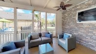 This single-floor space is thoughtfully designed for entertaining and relaxation, combining a fully equipped interior kitchen with a spacious screened outdoor lounge area. The kitchen is outfitted with rich dark cabinetry, dark granite countertops, stainless steel appliances including a refrigerator, microwave, and range, and a double basin sink. Decorative pendant lighting with warm-toned shades and a tropical leaf ceiling fan add character to the culinary space. A striking stacked stone accent wall frames the kitchen area, lending a natural, textured aesthetic that complements the dark cabinetry and granite surfaces. The bar-style counter with seating provides an inviting gathering point. The screened outdoor lounge area extends the living space seamlessly, offering panoramic views of lush, landscaped grounds with palm trees and manicured greenery. Beamed ceilings and another tropical ceiling fan enhance the airy, resort-like atmosphere. Comfortable wicker and rattan seating arrangements with blue accent cushions create a welcoming environment. Floor-to-ceiling screened panels flood the space with natural light while maintaining an open, breezy feel. Together, the interior and outdoor areas create a cohesive, amenity-rich environment ideal for communal use.