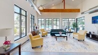 Located at 7515 Foxhall Ln in Henrico, Virginia, this single-story clubhouse delivers a striking blend of modern industrial and warm residential design. The space immediately impresses with its soaring vaulted ceilings, accented by bold exposed wooden beams and visible ductwork that lend the interior an open, loft-inspired character. Floor-to-ceiling black-framed windows line the walls, flooding the space with abundant natural light and providing panoramic views of the surrounding mature trees and an outdoor pool area.

The expansive main lounge serves as the heart of the space, featuring polished concrete floors, a large decorative area rug, and a comfortable seating arrangement with yellow armchairs and a teal sofa centered around a wooden coffee table. The inviting layout is ideal for both casual gatherings and relaxed socializing.

Adjacent to the lounge, a well-appointed kitchenette offers dark wood cabinetry, granite countertops with a curved island, stainless steel appliances including a full-size refrigerator and microwave, and a double sink — providing convenient amenities for residents and guests alike. Pendant lighting throughout adds warmth to the otherwise clean, contemporary aesthetic.

The overall ambiance is bright, airy, and welcoming, combining functional community spaces with thoughtful design details that create a comfortable and attractive environment.