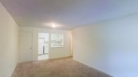 This single-floor space offers a clean, functional layout designed for comfortable everyday living. The living area is finished with wall-to-wall neutral-toned carpeting, creating a warm and inviting atmosphere. A textured ceiling and soft overhead lighting contribute to the room's understated, practical character. The living space flows openly toward the kitchen, which is visible through a pass-through opening that enhances the sense of connectivity between rooms. The kitchen features white cabinetry along the upper and lower walls, a white refrigerator, a stove, and a dishwasher, all arranged efficiently within a compact footprint. The light-colored tile flooring in the kitchen contrasts subtly with the carpeted living space, helping to visually define each functional zone. The walls throughout are painted in a neutral off-white tone, providing a bright, clean backdrop that can easily accommodate a variety of furnishing styles. A doorway adjacent to the kitchen suggests access to additional rooms, offering a straightforward and practical floor plan. Overall, the space presents a simple, well-maintained interior that is move-in ready and adaptable to a range of personal aesthetics.