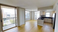 This single-floor space features an open-concept layout that seamlessly connects the living area and kitchen, creating a functional and spacious environment. Light hardwood flooring extends throughout, complementing the neutral beige walls and white trim. Large windows on two walls flood the interior with natural light while framing elevated city views, giving the space a distinctly urban character. A sliding glass door fitted with vertical blinds opens onto a private balcony, offering additional outdoor access and views of the surrounding cityscape. The kitchen is positioned at one end of the open plan, featuring dark countertops that contrast with the light cabinetry and flooring. Upper cabinets provide ample storage, and the peninsula-style counter acts as a natural divider between the kitchen and living areas. The living space is generously proportioned, with enough room to accommodate a variety of furniture arrangements. A textured ceiling and flush-mount lighting complete the interior. The overall ambiance is clean, modern, and light-filled, making it a practical and versatile urban space.