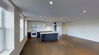 This single-floor space showcases a thoughtfully designed open-concept layout that seamlessly connects the kitchen and living areas. The kitchen is a standout feature, combining functionality with modern aesthetics. Upper cabinetry in a high-gloss white finish contrasts sharply with the darker lower cabinets, creating a striking two-tone effect. A full white subway tile backsplash extends across the cooking area, adding a clean, classic element to the contemporary design. The kitchen is equipped with white appliances, including a gas range and built-in microwave, lending a cohesive look to the space. A large central island with a light countertop and dark base serves as both a workspace and a social hub, illuminated by two pendant lights suspended from the ceiling. Recessed lighting throughout ensures the space is well-lit, complementing the natural light that filters in. Rich, dark hardwood flooring flows continuously from the kitchen into the open living area, grounding the space with warmth and texture. The expansive living area offers generous room for flexible furnishing arrangements, while the clean white walls and smooth ceilings contribute to a bright, minimalist ambiance overall.