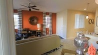 This two-floor space makes a strong visual impression with its thoughtfully designed living area. The main living space features a striking burnt-orange accent wall that serves as a bold focal point, complemented by warm yellow-toned walls that create an energetic yet cohesive color palette. A ceiling fan is centrally mounted overhead, providing both comfort and a practical design element. Natural light filters into the room through a window dressed with navy and white patterned curtains, adding a graphic contrast to the warm wall tones. The living area is furnished with a neutral-toned sofa and layered with a patterned area rug in navy and white, grounding the space. A decorative sunburst mirror mounted above a wooden storage credenza adds an artistic touch to the accent wall. Carpeted flooring runs throughout the main living area, contributing to a comfortable underfoot feel. A front entry door is visible within the living space, offering a sense of the overall layout. The open sightlines suggest a functional, well-connected floor plan that extends across two levels.