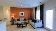 This single-floor space makes a strong visual impression through its thoughtfully designed living area. The room is anchored by a striking terracotta-orange accent wall, which adds warmth and depth to the overall aesthetic. Neutral beige tones on the remaining walls create a balanced contrast, ensuring the bold color does not overwhelm the space. Track lighting runs along the ceiling, providing flexible and well-distributed illumination throughout the room. Natural light filters in through a window dressed with elegant dark curtains, adding a touch of sophistication while allowing brightness into the interior. The living area is furnished with two light-blue sofas arranged around a dark wood oval coffee table, creating a comfortable and cohesive seating arrangement. Decorative pillows in earthy and metallic tones complement the color palette. A framed piece of wall art adds a refined focal point to the accent wall, while a decorative folding screen in the corner introduces texture and visual interest. Plush carpeting underfoot enhances the cozy atmosphere of the room. Overall, the space presents a harmonious blend of bold color, contemporary design, and comfortable furnishings, making it a welcoming and visually engaging environment.