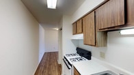 This single-floor residence offers a straightforward and functional layout across several well-defined spaces. The kitchen is a notable highlight, featuring an abundance of warm-toned wood cabinetry—both upper and lower—that provides generous storage throughout. White laminate countertops offer ample prep space, complemented by a double stainless steel sink, a gas range with overhead ventilation, and a dishwasher. Wood-look plank flooring runs through the kitchen and extends into the hallway, adding a cohesive and easy-to-maintain aesthetic.

A hallway connects the kitchen to the remaining rooms, with a louvered closet door visible along the corridor. The living area is spacious and carpeted in a deep brown tone, featuring a large sliding window that allows natural light to fill the room. Textured ceilings are found throughout the unit, a consistent architectural detail across all spaces.

The bedrooms are also carpeted and neutrally painted, providing clean, move-in ready conditions. One bedroom includes a built-in closet with louvered doors. The bathroom is accessed via a short hallway flanked by open shelving, and features wood-look flooring, a toilet, and a full tub and shower combination with a grab bar and curtain rod.
