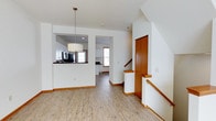 This three-floor home is designed with a clean, functional layout that balances open communal spaces with practical flow between levels. The main living area features light hardwood-style flooring that runs throughout, lending warmth and continuity to the space. A central open layout connects the living area to the kitchen, which is accessible via a pass-through counter with a dark surface, offering a convenient serving and social focal point. The kitchen is equipped with dark appliances, white cabinetry, and receives natural light through a double-hung window. A drum pendant light fixture anchors the living area visually. A staircase with warm wood handrails and white balusters provides access to the upper and lower floors, positioned conveniently near a storage closet with a wood-paneled door. The interior features white walls and warm wood trim throughout, creating a crisp yet inviting aesthetic. A private porch extends the living space outdoors, offering views of the surrounding neighborhood streetscape, complete with white railings and a wood deck floor — ideal for enjoying seasonal outdoor moments.