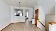 This three-floor home is designed with a clean, functional layout that balances open communal spaces with practical flow between levels. The main living area features light hardwood-style flooring that runs throughout, lending warmth and continuity to the space. A central open layout connects the living area to the kitchen, which is accessible via a pass-through counter with a dark surface, offering a convenient serving and social focal point. The kitchen is equipped with dark appliances, white cabinetry, and receives natural light through a double-hung window. A drum pendant light fixture anchors the living area visually. A staircase with warm wood handrails and white balusters provides access to the upper and lower floors, positioned conveniently near a storage closet with a wood-paneled door. The interior features white walls and warm wood trim throughout, creating a crisp yet inviting aesthetic. A private porch extends the living space outdoors, offering views of the surrounding neighborhood streetscape, complete with white railings and a wood deck floor — ideal for enjoying seasonal outdoor moments.