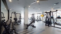 This well-appointed fitness center is designed to accommodate a comprehensive range of workout needs across a generously sized, single-level layout. The space is divided into several distinct functional zones, each thoughtfully equipped and finished to professional standards.

The main workout floor features a robust selection of strength training equipment, including cable machines, multi-use weight racks, barbells, and adjustable benches arranged across a durable striped rubberized floor. Large floor-to-ceiling windows line the perimeter, flooding the space with natural light and providing outdoor views while a series of ceiling fans maintain air circulation throughout.

A dedicated cardio zone houses ellipticals, treadmills, and stationary machines, many equipped with individual monitors. Full-length mirrors are mounted along several walls, enhancing the sense of openness. A separate glass-enclosed studio area with a warm terracotta ceiling accent provides space for functional training and group classes, outfitted with Echelon bikes, medicine balls, and a dumbbell rack. The studio connects visually to the main floor through floor-to-ceiling glass partitions with bold black framing. Recessed lighting throughout ensures consistent, bright illumination across all workout areas.