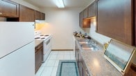 This single-floor unit presents a straightforward and functional layout designed for comfortable everyday living. The galley-style kitchen is efficiently arranged with dark espresso cabinetry lining both sides, laminate countertops, a double-basin stainless steel sink, and a white electric range, all set atop light-toned tile flooring that adds brightness to the space. The living area features plush wall-to-wall carpeting, neutral cream-toned walls, and a textured ceiling, creating a clean and understated atmosphere. A front entry area includes a closet with warm wood-toned doors, providing convenient storage just inside the entrance, with a partial view into the kitchen beyond. The bedroom spaces are each carpeted and equipped with baseboard heating units, windows dressed with horizontal blinds that allow for natural light, and a textured ceiling finish consistent throughout the unit. The bathroom is appointed with a granite-look vanity countertop, a toilet, and a tub-shower combination featuring a decorative floral shower curtain, complemented by tile flooring and a ventilation unit overhead. Overall, the unit maintains a clean, neutral aesthetic with warm wood accents on the doors and trim, making it a practical and move-in ready space.
