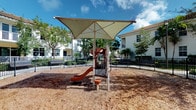 This outdoor community playground offers a well-designed recreational space for children, situated within a residential setting. The centerpiece of the area is a multi-function play structure equipped with a slide and climbing elements, all sheltered beneath a large, canopy-style shade sail supported by metal posts. The generous canopy provides meaningful sun protection, making the space usable even during bright, sunny days typical of a warm climate. The ground surface consists of wood chip mulch, providing a soft and impact-absorbing layer beneath the play equipment. The playground is enclosed by a sturdy black metal fence, creating a safe and defined boundary for the space. Mature trees and neatly maintained tropical landscaping line the perimeter, adding greenery and a sense of enclosure. The surrounding residential buildings, featuring tile rooflines and white facades, are visible beyond the fence, reinforcing the community-oriented nature of this amenity. The overall atmosphere is bright, open, and inviting, offering a dedicated and thoughtfully equipped outdoor space well-suited for active play within a neighborhood setting.