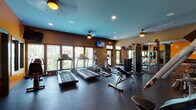 This single-floor fitness center is thoughtfully designed to support a full range of workout activities. The main exercise area is spacious and open, featuring durable rubber flooring throughout to accommodate heavy equipment and high-impact training. A row of treadmills and elliptical trainers lines the windowed wall, where large windows with shutters allow ample natural light to flood the space and provide views of the surrounding outdoor environment. Multiple ceiling fans are mounted above to maintain air circulation during workouts.

The strength training section is well-appointed with a multi-station cable machine, a dedicated dumbbell rack with a full set of free weights, adjustable benches, and additional resistance machines for targeted muscle training. Full-length mirrors on one wall enhance the sense of space and allow users to monitor their form. Wall-mounted flat-screen televisions are positioned throughout for entertainment during cardio sessions.

A bold orange accent wall adds a vibrant, energizing focal point to the strength training zone, complementing the blue-painted ceiling that gives the space a distinctive, modern aesthetic. An adjoining children's activity room is accessible from the main gym floor, furnished with a table, chairs, a wall-mounted television, and playful décor, making it convenient for parents to work out while keeping young children nearby.