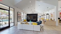 Located at 9111 Lakes at 610 Dr in Houston, TX, this single-story clubhouse is a dynamic community gathering space defined by its contemporary design and energetic aesthetic. Soaring vaulted ceilings with abundant recessed lighting create an open, airy atmosphere throughout, while large floor-to-ceiling windows and glass doors flood the interior with natural light and frame views of the resort-style pool and surrounding landscaping.

The main lounge area anchors the space with a generous white sectional sofa, bold yellow accent pillows, and a large wall-mounted flat-screen TV set against a dramatic dark feature wall. Vibrant framed artwork and eye-catching pop-art wall graphics inject personality and color throughout. Light wood-look flooring and large-format tile flow seamlessly across the space, complementing the clean, white walls.

A dedicated co-working zone features bar-height counters, task seating, and stylish pendant lighting, providing a functional yet inviting workspace. Booth-style lounge seating with patterned upholstery and yellow cushions offers additional casual gathering spots. A fully equipped kitchen with dark cabinetry, white quartz countertops, stainless steel appliances, and a large island opens directly toward the pool area through expansive glazing. A cheerful yellow café nook near the entrance offers a bright, intimate spot with direct pool access.