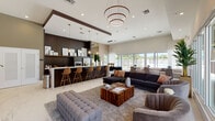 Located in Miami, FL, this single-story clubhouse at SW 152nd Street serves as a sophisticated community hub designed for both socializing and relaxation. The open-plan layout is anchored by polished marble-look tile flooring that flows seamlessly throughout, reflecting abundant natural light pouring in through floor-to-ceiling windows dressed in sheer white drapery.

The central lounge area is generously appointed with plush sectional sofas, tufted seating, and richly grained wood coffee tables, all beneath a striking multi-ring copper chandelier that serves as a bold focal point. Adjacent to the lounge, a sleek bar counter with leather-upholstered barstools connects to a fully outfitted kitchen featuring dark wood cabinetry, a stainless steel refrigerator, microwave, and pendant globe lighting — ideal for catered events or casual gatherings.

A dedicated dining nook with bistro-style tables and upholstered chairs provides an intimate setting for smaller groups, while glass-panel walls offer views into a fitness area visible from the main lounge. The overall design palette balances warm espresso tones against crisp white surfaces, creating a contemporary yet inviting ambiance that reflects the vibrant Miami lifestyle.