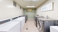 Located at 820 Capital Dr in Carnegie, PA, this community laundry facility offers a practical and well-maintained space designed to accommodate residents with ease. The single-floor layout is thoughtfully organized, encompassing two dedicated laundry rooms and a shared waiting area.

Each laundry room is outfitted with multiple top-loading washers and front-loading washer-dryer units, arranged in parallel rows to maximize efficiency. The rooms feature light wood-look flooring, painted concrete block walls in a soft sage green, and bright overhead fluorescent lighting that ensures excellent visibility. Ventilation is provided through wall-mounted vents, and a utility sink is accessible within the laundry area.

The waiting area provides a comfortable space for residents during their laundry cycles. It is furnished with sleek, black multi-seat benches and includes a work surface for convenience. Decorative wall art adds a welcoming touch, while a vending machine offers refreshments. The waiting area also benefits from a skylight and overhead lighting, creating a bright and pleasant atmosphere. A restroom is accessible directly from this space.

Throughout the facility, the consistent use of light flooring and sage-toned walls creates a cohesive, clean aesthetic. The layout is straightforward and functional, making this laundry facility a reliable and comfortable shared amenity.