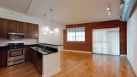 The Kahlo is a well-appointed 1-bedroom, 1-bathroom residence with a dedicated study, offering a thoughtfully designed single-floor layout that balances comfort and functionality. The open-concept living and kitchen area anchors the space, showcasing warm hardwood flooring that runs throughout and adds a timeless character to the interior. A striking exposed brick accent wall serves as a defining architectural feature, lending an industrial-chic aesthetic that contrasts beautifully with the clean white walls and ceilings. The kitchen is equipped with dark countertops and rich wood cabinetry, complemented by a peninsula with an integrated sink — ideal for both meal prep and casual dining. Stylish pendant lighting hangs above, providing focused illumination while enhancing the overall design. Natural light flows generously into the living area through a large window fitted with blinds, while a full-height sliding glass door with vertical blinds offers access to an outdoor area and further brightens the space. The study provides a versatile additional room suitable as a home office or flex space. Recessed lighting throughout ensures consistent, even illumination. The overall ambiance is modern and inviting, blending industrial elements with warm, natural materials.