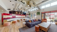 This dynamic clubhouse is a thoughtfully designed social hub that combines modern aesthetics with a lively, energetic atmosphere. The space is anchored by soaring vaulted ceilings accented with exposed white-painted beams and bold black structural elements, creating an airy, open feel throughout. Statement pendant light fixtures in sculptural gold wire designs hang overhead, adding an artistic touch to the interior.

The main lounge area features a generously sized tufted charcoal sectional sofa adorned with colorful accent pillows, surrounded by natural wood stump stools, wire side tables, and eclectic woven ottomans. A standout curved built-in bench, framed by warm natural wood and vibrant red paneling, serves as both a focal point and flexible gathering spot. A ping-pong table sits centrally, reinforcing the space's recreational character.

Bold, large-scale artwork is displayed against a textured white stone accent wall, complemented by lush trailing greenery suspended from the upper walls, bringing nature indoors. Light wood-plank flooring flows throughout, enhancing the warm, welcoming ambiance.

Large black-framed sliding glass doors and expansive windows flood the interior with natural light and frame views of a covered outdoor patio with dining seating, a pool, and surrounding landscaping. The seamless indoor-outdoor connection makes this clubhouse an exceptional space for gathering, recreation, and relaxation.