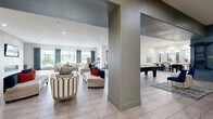 This expansive single-floor community clubhouse offers a generously proportioned, open-concept layout designed for both relaxation and socializing. Large format tile flooring extends throughout, anchoring the space with a clean, contemporary aesthetic, while structural columns define distinct activity zones without compromising the open feel.

The main lounge area features multiple seating arrangements, including stylish striped accent chairs and plush sofas, centered around a herringbone-patterned area rug. A wall-mounted flat-screen television and a sleek linear fireplace serve as a refined focal point, complemented by floor-to-ceiling windows dressed in bold geometric-patterned drapes that frame unobstructed views of the outdoor pool and landscaped courtyard.

Adjacent to the lounge, a recreation zone houses a full-size billiards table, providing an entertaining gathering spot. The dining area features multiple seating options, including a bar-height counter with elegant pendant globe lighting above and dark tile accents, as well as a separate dining table surrounded by rich blue velvet chairs. A vaulted ceiling adds vertical drama to this section of the space.

Additional amenities include a package locker system, workstation areas, and decorative wall art that adds personality throughout. Recessed lighting and abundant natural light from expansive windows create a bright, welcoming ambiance suited for community gatherings.