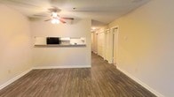 This single-floor residence offers a clean, functional layout designed for comfortable everyday living. The main living area is finished with rich, wood-toned laminate flooring that extends throughout the space, lending warmth and visual continuity. A ceiling fan with an integrated light fixture is centrally mounted, providing both air circulation and ambient illumination.

The kitchen is positioned adjacent to the living area and features a pass-through counter, creating an open connection between the two spaces. White cabinetry provides ample storage, while a black appliance suite, including a refrigerator and range, offers a practical contrast to the light-colored walls and cabinetry.

A hallway extends from the main living area, lined with multiple doors that lead to additional rooms. The hallway features louvered bi-fold closet doors, suggesting generous storage options throughout. The walls are painted in a neutral warm beige tone, creating a bright and welcoming atmosphere.

The textured ceiling adds a subtle architectural detail consistent throughout the space. Overall, the residence presents a straightforward and well-maintained interior that balances open communal areas with defined private spaces, making it a practical and inviting environment.