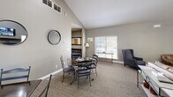 This single-floor communal space is thoughtfully designed to offer both comfort and functionality across several interconnected areas. The living room serves as the heart of the space, featuring soaring vaulted ceilings that lend an open, airy feel. A stone-surround fireplace with a rich wood mantel anchors one wall, complemented by a wall-mounted flat-screen TV, creating an inviting focal point. Comfortable leather seating and plush upholstered wingback chairs are arranged throughout, providing ample seating for gatherings. Natural light streams in through multiple windows dressed with classic blinds, brightening the neutral gray walls and patterned carpeted floors. The dining area transitions seamlessly from the living space, accommodating multiple dining tables and chairs beneath the continued vaulted ceiling. Decorative round mirrors add a stylish touch to the walls in this section. Beyond the dining area, a fully equipped kitchen is visible, featuring stainless steel appliances, dark cabinetry, a refrigerator, and bar-height seating. The open-concept layout encourages easy flow between zones, making this space well-suited for socializing, dining, and relaxing. A glimpse into an adjacent room suggests additional recreational or activity space, rounding out this versatile and well-appointed interior.
