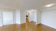 This single-floor space offers a clean, functional layout designed for comfortable everyday living. The main living area is open and airy, finished with warm-toned hardwood-style flooring that runs continuously throughout, creating a cohesive and inviting atmosphere. Crisp white walls and a smooth white ceiling reflect natural and artificial light effectively, enhancing the sense of openness across the space.

From the living area, a wide doorway leads directly into the kitchen, which is visible in the background and features white cabinetry and dark countertops, providing a practical and tidy workspace. Adjacent to the kitchen entry, a paneled white door with a classic six-panel design leads to an additional room, likely a closet or utility space, contributing to the unit's overall storage potential.

The layout flows naturally, with clear sightlines from the main living space toward the kitchen and connecting areas. Flush-mounted ceiling lighting provides consistent illumination throughout. The overall ambiance is neutral, clean, and move-in ready, making it a versatile space well-suited to a variety of furnishing styles and personal preferences.