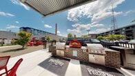 Located at 1308 Roseneath Rd in Richmond's vibrant Scott's Addition neighborhood, this single-story amenity space at The Preserve combines industrial-chic design with modern social features. The interior showcases exposed brick walls, warm wood-paneled ceilings, and expansive floor-to-ceiling steel-framed windows that flood the space with natural light while offering views of the surrounding urban streetscape. A full-size billiards table anchors the game area, complemented by high-top bar tables with stylish seating and comfortable lounge furniture arranged throughout. Bold branded artwork on the white brick walls adds a distinctive local character to the interior. Large glass doors open directly to a covered outdoor patio, seamlessly connecting the inside and outside areas. The exterior entertaining zone is well-equipped with a built-in brick grilling station featuring multiple stainless steel grills and a standout wood-fired pizza oven as a focal point. Red Adirondack chairs and café-style tables and chairs complete the outdoor seating area beneath a corrugated metal pergola, creating a versatile space for communal gatherings and outdoor cooking in the heart of one of Richmond's most dynamic neighborhoods.