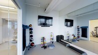 This single-floor fitness space is designed to support a wide range of exercise activities within a clean, minimalist setting. The room features white walls and a light-toned floor, creating a bright and open atmosphere that feels larger than its footprint. A large wall-mounted flat-screen television is positioned at an ideal viewing height, accompanied by a soundbar, allowing users to follow workout classes or entertainment during training sessions. A full-length mirror spans one wall, enhancing the sense of space while enabling users to monitor their form during exercise. The room is well-stocked with functional training equipment, including a rack of medicine balls in various sizes, a tiered dumbbell stand, kettlebells arranged on a dedicated rack, weight plates stacked neatly, and a BOSU balance trainer. A treadmill is visible along one side, catering to cardiovascular training needs. Rolled yoga or exercise mats are stored upright, and additional small equipment pieces are organized throughout. Exposed ceiling elements and panel-style walls add an industrial character to the otherwise streamlined space, making it a practical and efficient private or shared fitness facility.