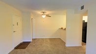This single-floor residential space offers a clean, functional layout designed for comfortable everyday living. The main living area is finished with wall-to-wall carpeting in a warm neutral tone, providing a cozy and inviting atmosphere. Crisp white walls and white trim throughout lend the space a bright, well-maintained appearance. A ceiling fan with an integrated light fixture is centrally mounted overhead, offering both ambient lighting and air circulation year-round. A transitional area near the entry features a hard flooring surface, creating a practical separation from the carpeted living space. A pass-through counter opening connects the living area to an adjacent space, enhancing the open feel and allowing for easy interaction between rooms. An interior door leads to additional areas of the home, contributing to the overall flow of the layout. Neutral tones dominate the color palette, making the space highly versatile for a variety of furnishing styles. The overall ambiance is understated and move-in ready, with a straightforward design that prioritizes livability and practicality.