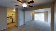 This single-story space features an open layout that connects the main living area to the kitchen. The living room is finished with wall-to-wall carpeting and neutral-toned walls, creating a clean and understated backdrop. A ceiling fan with a built-in light fixture is centrally mounted, providing both air circulation and ambient lighting throughout the room. A large sliding glass door spans much of the far wall, fitted with vertical blinds that allow for adjustable natural light and potential outdoor access. The space includes multiple interior doors, offering access to additional rooms within the unit. Adjacent to the living area, the kitchen features tile flooring that contrasts with the carpeted living space, and includes a full-size refrigerator. The transition between the two areas is clearly defined by the flooring change, giving each zone a distinct character while maintaining an overall sense of openness. The textured ceiling and straightforward finishes throughout reflect a practical, functional design.