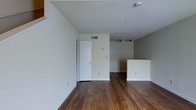 This two-story space showcases a clean, functional layout with warm, dark hardwood flooring that runs throughout the main level, adding richness and character to the interior. The living area is open and airy, defined by neutral white walls and a textured popcorn ceiling that adds subtle depth overhead. A set of track lights is mounted near the ceiling, providing adjustable, directed illumination across the space — a practical and stylish lighting solution.

A white kitchen peninsula is visible toward the rear of the main living area, offering a natural division between the living and kitchen zones while maintaining an open-concept feel. The peninsula provides additional counter space and casual seating potential. A ceiling-mounted HVAC vent ensures climate control is integrated discreetly into the layout.

A doorway with a standard white door offers access to an adjacent room or closet, contributing to the overall flow of the floor plan. The walls are clean and move-in ready, painted in a soft, neutral tone that complements the warm flooring. The overall ambiance is understated and versatile, lending itself well to a variety of furnishing styles across its two levels.