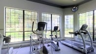 This fitness center is designed to provide a functional and inviting space for exercise and wellness. The room is anchored by a row of large, grid-pane windows that span the exterior wall, flooding the space with natural light and offering unobstructed views of the surrounding green landscape. The bright, airy atmosphere makes for an energizing workout environment throughout the day.

The cardio area is well-appointed with a selection of exercise equipment, including elliptical trainers, an upright stationary bike, and a treadmill, all arranged to take full advantage of the garden views. The neutral-toned walls and light flooring contribute to a clean, modern aesthetic that keeps the space feeling open and uncluttered.

A wall-mounted security or display monitor is visible in the upper corner of the room, reflecting attention to both safety and convenience. The dark crown molding along the upper perimeter of the room adds a subtle architectural detail that frames the space with a polished finish. Overall, the fitness center balances practicality with a welcoming ambiance, offering a comfortable setting for a range of cardio-focused workouts in a naturally lit, visually open environment.
