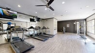 Located at 11245 West Rd in Houston, Texas, this well-equipped fitness center offers a dedicated exercise room designed to support a well-rounded workout routine. The space features light wood-style flooring throughout, lending a clean and modern aesthetic that complements the neutral gray and white color palette of the walls.

Cardio equipment includes multiple treadmills and an elliptical trainer, positioned in front of a large wall-mounted mirror that visually expands the space and allows users to monitor their form. A mounted flat-screen TV above the mirror adds an entertainment element to cardio sessions. A ceiling fan provides additional air circulation overhead, complemented by recessed lighting that ensures the room is consistently well-lit.

For strength training, the room is outfitted with a multi-function cable machine, an adjustable workout bench, and a tiered dumbbell rack stocked with a range of free weights. A collection of stability and medicine balls rounds out the functional fitness offerings, supporting flexibility and core training.

Natural light filters in through multiple windows, and a set of glass-paneled French doors provides both natural illumination and easy access. The open floor plan allows ample space for stretching or floor exercises, making this fitness center a comprehensive and functional facility for residents.