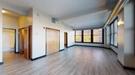 Located along SR-2 E in Cleveland, OH, this single-floor residence offers an urban living experience with a thoughtfully designed layout and contemporary finishes. Light wood-toned luxury vinyl plank flooring flows consistently throughout the space, complemented by crisp white walls and bold dark baseboards that define each room with clean contrast.

The open main living area is a standout feature, characterized by an impressive bank of large, dark-framed windows that frame sweeping views of the Cleveland cityscape and surrounding brick architecture. Exposed industrial ductwork adds an urban loft character to the space, while abundant natural light fills the interior throughout the day.

The kitchen is generously proportioned, featuring warm honey-toned wood cabinetry, dark granite countertops, and black appliances including a range, microwave, refrigerator, and dishwasher. A peninsula with a pendant light overhead provides a natural transition between the kitchen and the living area.

Additional rooms include a well-sized bedroom with its own HVAC vent and recessed ceiling detail, a dedicated closet with double wood-panel doors, and a full bathroom equipped with a tub/shower combination, wood vanity with granite countertop, and light wood-look tile flooring. A second full bathroom mirrors this clean, functional aesthetic. The overall ambiance is modern, bright, and urban.