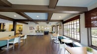 Located at 605 Hampshire Rd in Thousand Oaks, CA, this single-story community clubhouse offers a well-appointed gathering space designed for socializing and recreation. The main lounge area is defined by striking exposed beam ceilings with recessed skylight panels, dark hardwood-style flooring, and an open layout that promotes a sense of spaciousness throughout.

Large grid-pane windows dressed with woven bamboo shades line one wall, flooding the interior with natural light while offering views of the surrounding landscaping. Casual dining is accommodated through a mix of round and rectangular tables paired with modern chairs and teal booth-style seating. A billiard table anchors the game zone, adding a recreational focal point to the space.

The full kitchen features white cabinetry, stone-look countertops, stainless steel appliances including a refrigerator, electric range, and microwave, and a generous bar-height counter with seating for multiple guests, illuminated by contemporary pendant lighting. Patterned accent wallpaper adds visual interest to this area.

A wall of sliding glass doors opens directly to an outdoor patio equipped with a built-in grill and seating area, seamlessly extending the entertaining space outdoors against a backdrop of mature trees and California hills.