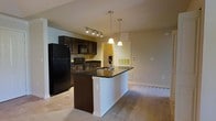 This single-floor residence offers a thoughtfully designed open-concept layout that seamlessly connects the kitchen and living areas. The kitchen serves as the heart of the space, showcasing rich dark wood cabinetry that contrasts elegantly with the dark granite countertops. A central island with a built-in sink provides additional workspace and casual dining potential, illuminated by two stylish pendant lights that hang above. Black appliances, including a full-size refrigerator, microwave, and range, complete the kitchen's cohesive aesthetic. A track lighting fixture overhead ensures ample task lighting throughout the cooking area. The transition from tile flooring in the kitchen to plush carpeting in the adjoining living area creates a natural delineation between the two spaces while maintaining an open, airy feel. Neutral-toned walls throughout allow for versatile decorating possibilities. The living area is generously proportioned, offering ample room for comfortable furnishings. Overall, the space strikes a balance between functionality and modern style, making it a practical yet visually appealing environment suitable for everyday living and entertaining.