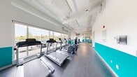 This modern fitness center offers a thoughtfully designed, single-floor layout with generous open floor space to accommodate a full range of exercise equipment. The facility is defined by its bright, airy atmosphere, courtesy of large floor-to-ceiling windows along multiple walls that flood the interior with natural light and provide views of the surrounding neighborhood. The clean white walls are accented by a bold teal horizontal band, giving the space a contemporary and energetic aesthetic that is reinforced by a prominent dumbbell logo graphic on the wall.

The cardio zone lines one side of the facility and features a row of treadmills and elliptical machines by Life Fitness, all positioned to face the windows for an engaging workout experience. The strength training area is equally well-appointed, with a cable machine, a power rack loaded with weight plates, a selection of benches, and a full dumbbell rack offering a range of weights. Large mirrors are strategically placed throughout, enhancing the sense of space and supporting proper form during exercise.

Underfoot, durable rubber and tile flooring provides safety and cushioning across the workout zones. The exposed ceiling incorporates visible ductwork and linear LED lighting, contributing to the facility's clean, industrial-modern character. The overall layout is open and unobstructed, creating a functional and motivating environment for a complete workout.