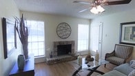 This single-story residence centers around a welcoming living area that blends functional design with comfortable character. The focal point of the room is a traditional brick fireplace with a raised hearth, adding warmth and a rustic charm to the space. Flanking the fireplace are multiple windows fitted with blinds, allowing ample natural light to fill the room while providing privacy control. The walls are painted in a soft, neutral tone that complements the warm hardwood-style flooring running throughout the living area. A textured popcorn ceiling adds visual depth overhead, while a ceiling fan with a light fixture ensures year-round comfort and illumination. Decorative touches, including a circular metal wall art piece above the fireplace mantel, enhance the room's aesthetic without overwhelming the space. A glass-top dining table is partially visible, suggesting a nearby dining area that flows naturally from the living space. A white exterior door positioned to the right of the fireplace offers convenient access to the outdoors, contributing to the open and airy feel of this single-level home.