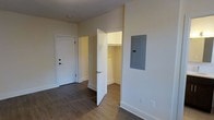 This single-floor space presents a straightforward, functional layout finished in a clean, neutral palette. The walls are painted in a soft off-white tone, complemented by crisp white baseboards and trim throughout, creating a bright and cohesive aesthetic. The flooring consists of dark hardwood-style planks, offering a warm contrast to the light walls and adding durability underfoot.

A notable feature of this space is the walk-in closet, accessible through a white paneled door. The closet is equipped with a hanging rod and benefits from its own interior lighting, providing practical and organized storage. Adjacent to the closet, an electrical panel is conveniently recessed into the wall, keeping utilities accessible while maintaining a tidy appearance.

The exterior entry door is solid and features a modern lever-style handle with a deadbolt, contributing to the security and functionality of the space. The overall finish quality appears fresh and well-maintained, suggesting recent construction or renovation. The combination of neutral finishes, durable flooring, and thoughtful storage solutions makes this a practical and adaptable space.