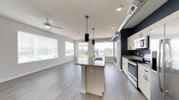 This single-floor space showcases a thoughtfully designed open-concept layout that seamlessly integrates the kitchen and living area. The kitchen is a standout feature, centered around a spacious island with a dark stone countertop and an undermount sink, complemented by two sleek pendant lights overhead. Light wood cabinetry pairs with stainless steel appliances, including a range and built-in microwave, while an exposed metallic ductwork detail adds an industrial accent against a bold dark wall. The living area extends generously from the kitchen, finished with light wood-look flooring that runs throughout, contributing to a clean and cohesive aesthetic. A ceiling fan provides both function and style overhead. Natural light pours in abundantly through multiple large windows fitted with white blinds, offering views of the surrounding cityscape and greenery. The overall ambiance is bright, airy, and contemporary, combining modern design elements with practical functionality to create a welcoming and versatile living environment.
